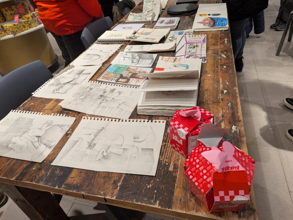 Sketchbooks spread across a table at Lush; group members are shown below the shoulders